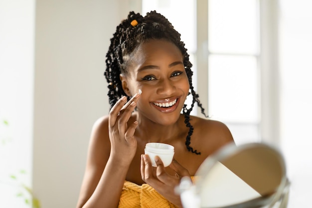 Foto retrato de uma linda mulher negra aplicando creme facial e uma senhora sorridente hidratando a pele com creme nutritivo para o dia