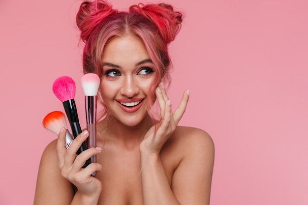 Retrato de uma linda mulher sem camisa com um penteado colorido segurando pincéis de maquiagem