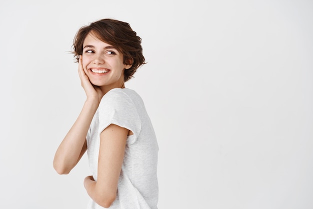 Retrato de uma linda mulher sincera com cabelo curto olhando para trás no espaço vazio olhando para a direita e sorrindo feliz em pé na camiseta no fundo branco