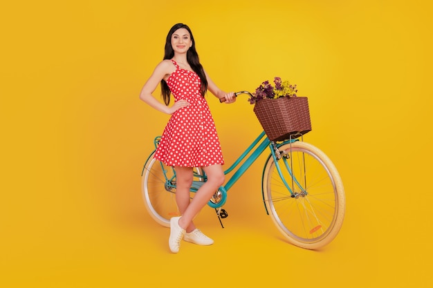 Retrato de uma linda senhora deslumbrante com uma bicicleta entregando flores e vestindo um minivestido vermelho pontilhado sobre fundo amarelo