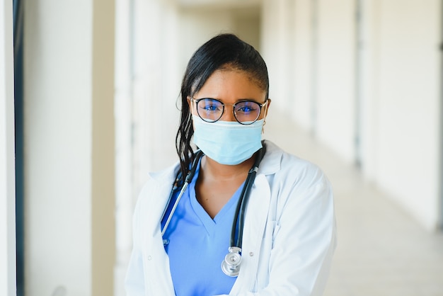 Retrato de uma médica afro-americana usando máscara em pé no corredor do hospital. medicina, saúde e serviços de saúde durante a pandemia de coronavírus covid 19.