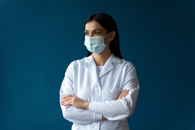 Retrato de uma médica atraente e séria usando máscara protetora, jaleco branco e estetoscópio e posando de braços cruzados fundo de cor azul isolado