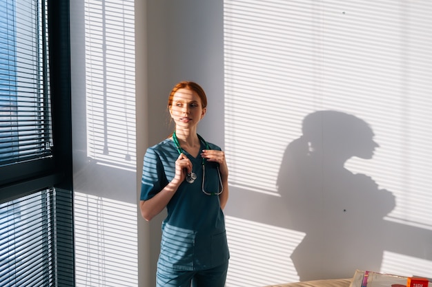 Retrato de uma médica com uniforme verde azul em pé perto da janela em um dia ensolarado no consultório da clínica