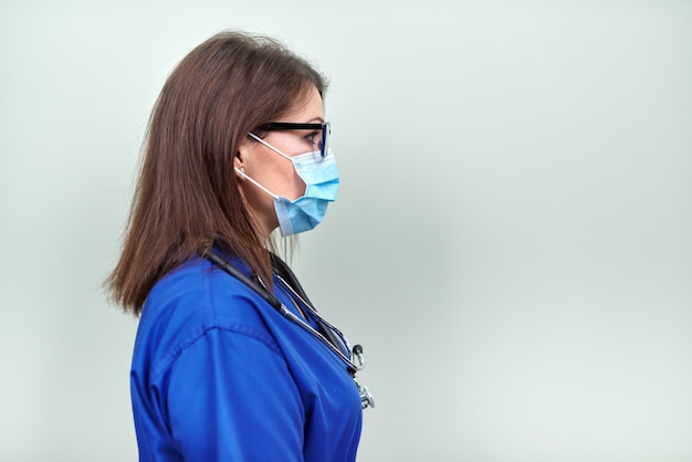 Retrato de uma médica confiante com máscara protetora médica de estetoscópio em uniforme azul, olhando de perfil em fundo verde pastel, copie o espaço