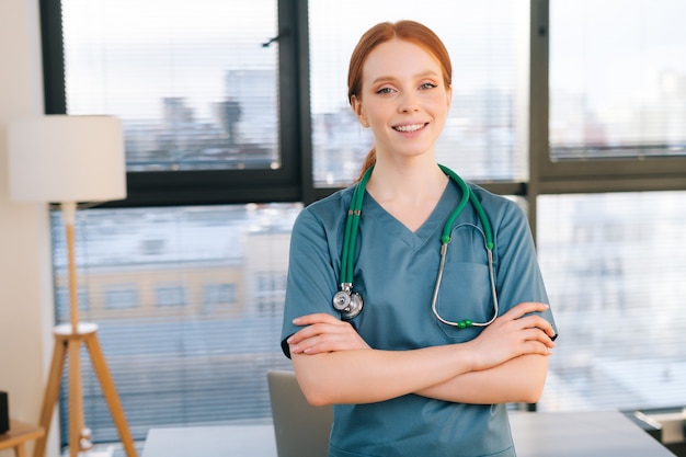 Retrato de uma médica simpática em uniforme verde azul em pé no fundo da janela em um dia ensolarado no escritório da clínica médica.