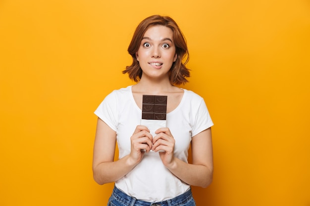 Retrato de uma menina adorável e feliz em pé, isolado na parede amarela, segurando uma barra de chocolate