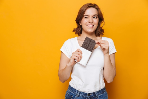 Retrato de uma menina adorável e feliz em pé, isolado na parede amarela, segurando uma barra de chocolate