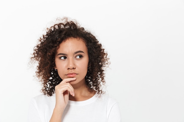 Foto retrato de uma menina africana casual muito pensativa em pé, isolada sobre uma parede branca, pensando