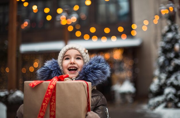 Retrato de uma menina alegre com uma caixa de presente para o Natal em uma rua da cidade no inverno com neve em um mercado festivo com decorações e luzes de fadas. Roupas quentes, chapéu de malha, lenço e pele. Ano Novo