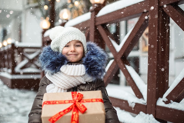 Retrato de uma menina alegre com uma caixa de presente para o Natal em uma rua da cidade no inverno com neve em um mercado festivo com enfeites e luzes de fada Roupas quentes chapéu de malha cachecol e peles ano novo