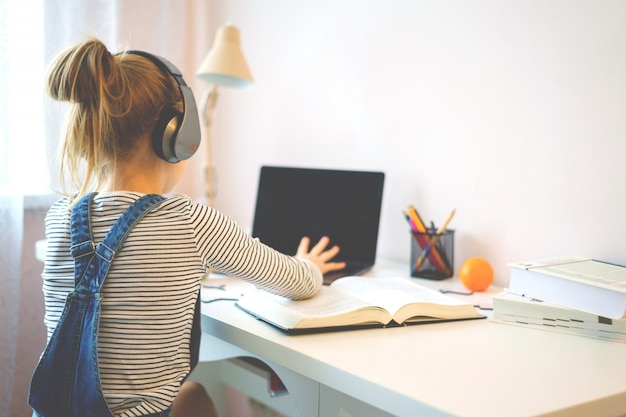Retrato de uma menina aprendendo on-line com fones de ouvido e laptop, anotando em um caderno, sentado em sua mesa em casa fazendo lição de casa