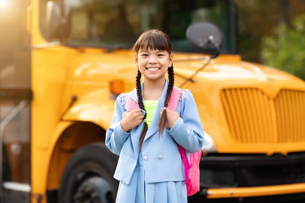 Retrato de uma menina asiática sorridente e feliz posando além do ônibus escolar amarelo ao ar livre