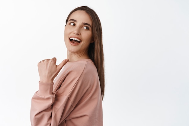 Retrato de uma menina bonita alegre apontando o polegar e olhando por trás do ombro, rindo e sorrindo enquanto olha no branco