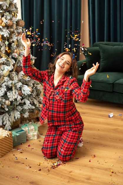 Retrato de uma menina bonita alegre de pijama, sentado perto da árvore de Natal sob chuva de confete e comemorando.