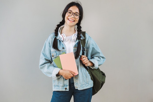 Retrato de uma menina bonita alegre em jaqueta jeans, usando óculos segurando a mochila e olhando para a câmera isolada sobre a parede cinza segurando livros.