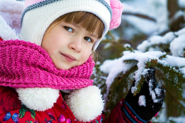 Retrato de uma menina bonita com roupas de inverno perto de um pinheiro coberto de neve