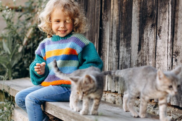 Retrato de uma menina bonita e feliz encaracolada com uma camisola de malha brincando com um gatinho perto de uma parede de madeira