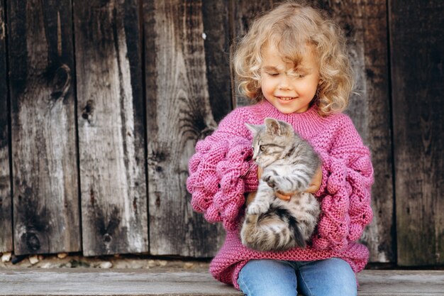 Retrato de uma menina bonita e feliz encaracolada com uma camisola de malha brincando com um gatinho perto de uma parede de madeira