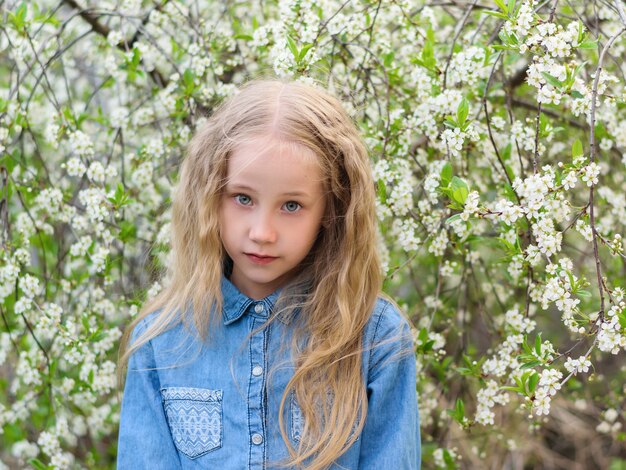 Retrato de uma menina bonita em uma camisa jeans com uma expressão facial séria em um pomar de cereja.