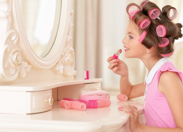 Retrato de uma menina bonita feliz com rolos de cabelo e batom perto do espelho