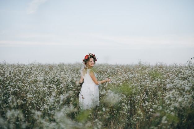 Retrato de uma menina bonita loira de olhos azuis, com uma coroa de flores na cabeça, andando em campo com flores brancas
