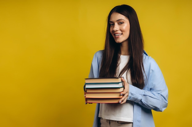 Retrato de uma menina bonita sorridente segurando livros e olhando para câmera isolada sobre fundo amarelo