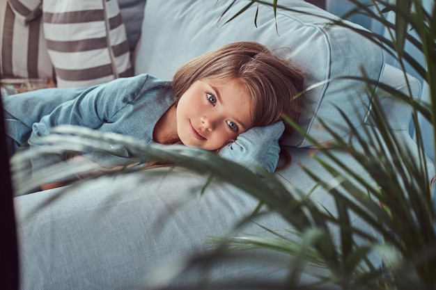 Retrato de uma menina bonitinha com longos cabelos castanhos e olhar penetrante, deitada em um sofá em casa sozinha.
