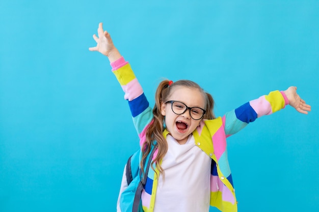 Retrato de uma menina bonitinha com óculos em uma jaqueta listrada com notebooks e uma mochila regozijando e se divertindo O conceito de educação Foto studio lugar de fundo azul para texto