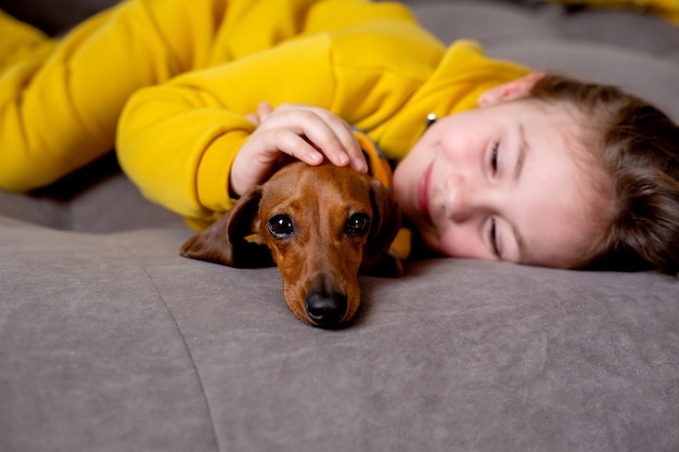 Retrato de uma menina bonitinha com roupas amarelas deitada na cama com um dachshund anão em macacão azul