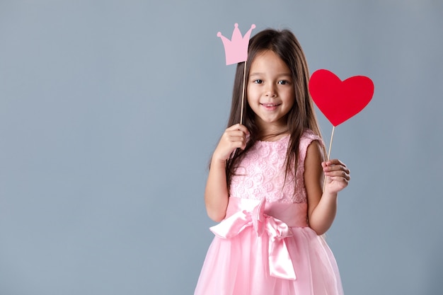 Retrato de uma menina bonitinha em um vestido rosa segurando um coração de papel e uma coroa de princesa