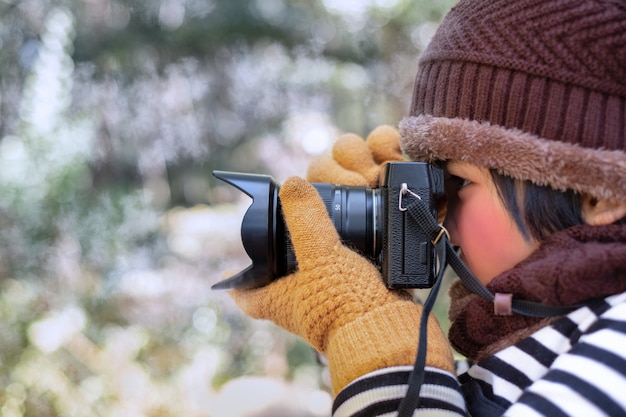 Retrato de uma menina bonitinha no chapéu de lã, tirando uma foto com a câmera digital no tempo está frio no fundo desfocado