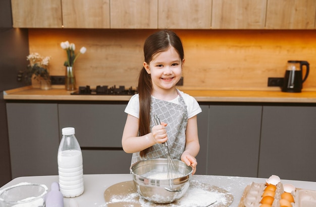 Retrato de uma menina bonitinha parada em uma cozinha moderna e preparando massa