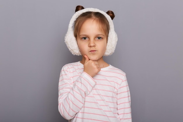 Retrato de uma menina bonitinha pensativa usando protetores de ouvido de pele e camisa listrada em pé isolado sobre fundo cinza em pé e segurando o queixo pensando em lições
