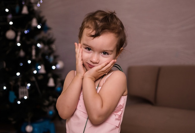 Retrato de uma menina branca com um vestido rosa em uma sala no contexto de uma árvore de natal