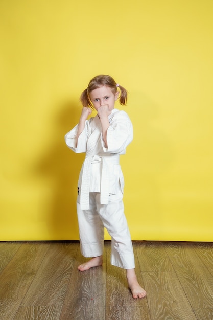 Retrato de uma menina caucasiana de 5 anos de quimono praticando karatê contra um fundo amarelo em casa