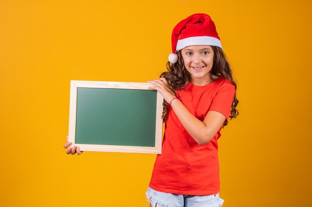 Retrato de uma menina caucasiana satisfeita com chapéu de Natal, segurando um banner com espaço para texto.