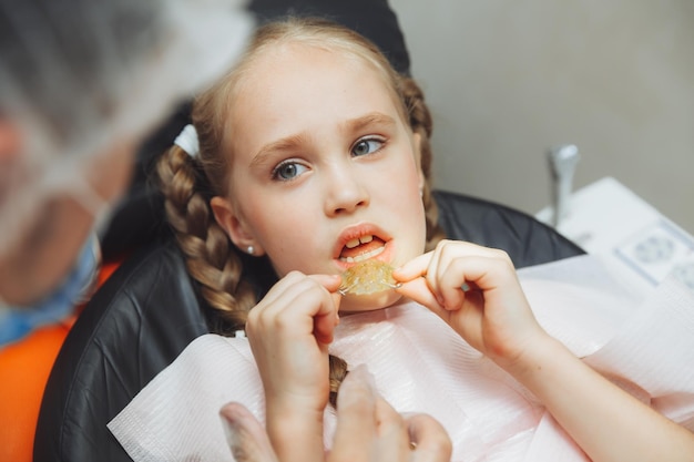 Retrato de uma menina com a boca aberta, sentado na cadeira de um dentista enquanto um ortodontista segura um prato nos dentes O dentista coloca um prato na boca de uma criança
