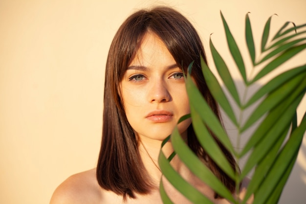 Retrato de uma menina com beleza natural, segurando um galho verde perto do rosto em fundo bege