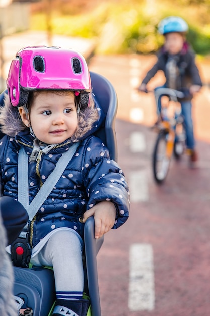 Retrato de uma menina com capacete de segurança na cabeça, sentada no assento da bicicleta e o irmão dela com a bicicleta