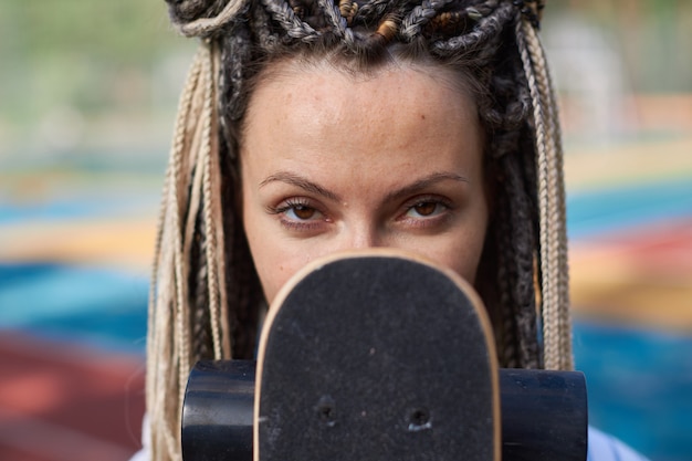 Retrato de uma menina com dreads que cobre o rosto com um longboard e olha diretamente para a câmera esporte moderno foto de alta qualidade