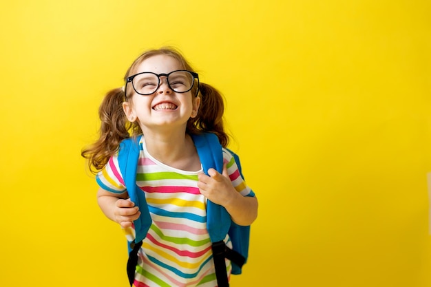 Foto retrato de uma menina com óculos e uma camiseta listrada com uma mochila escolar em um fundo amarelo