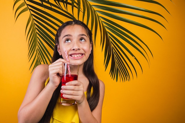 Retrato de uma menina com olhar travesso, segurando um suco de fruta em fundo amarelo.