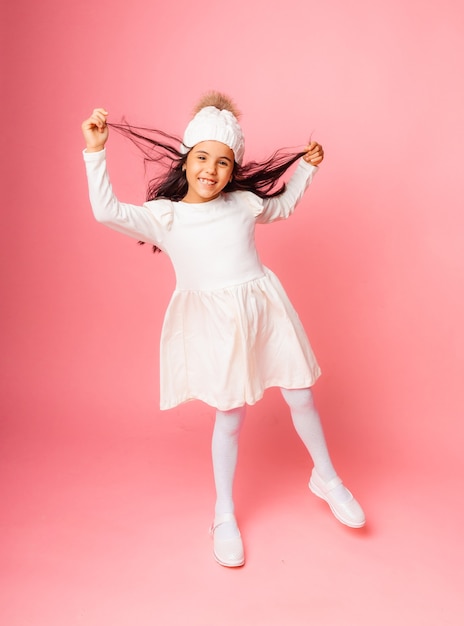 Retrato de uma menina com um chapéu branco de malha de inverno e um vestido branco em um fundo rosa