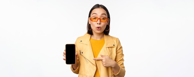 Retrato de uma menina coreana estilosa em óculos de sol sorrindo apontando o dedo para a tela do smartphone mostrando o aplicativo de celular sobre fundo branco