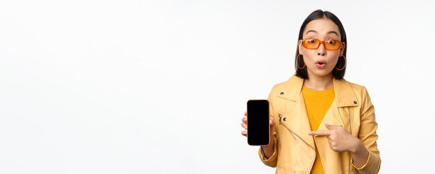 Retrato de uma menina coreana estilosa em óculos de sol sorrindo apontando o dedo para a tela do smartphone mostrando o aplicativo de celular sobre fundo branco