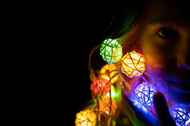 Foto retrato de uma menina criança de sete anos segurando uma guirlanda nas mãos perto do rosto, brilhando com luzes de néon multicoloridas coloridas durante a noite em casa. celebração do feriado de véspera de natal