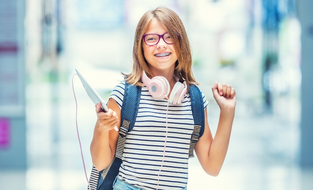 Retrato de uma menina da escola adolescente feliz moderna com mochila bolsa e fones de ouvido. Menina com aparelho dentário