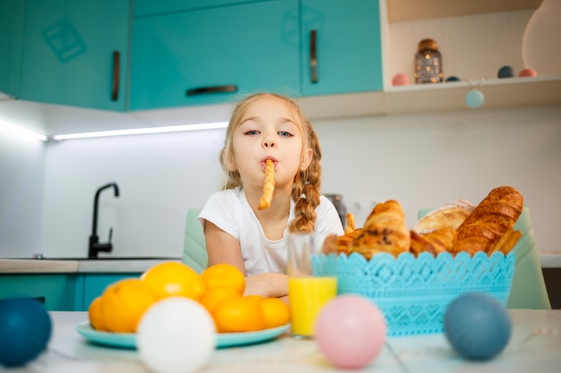 Retrato de uma menina de 7 anos de idade se senta na cozinha e brinca com palitos de pão. Criança toma café da manhã na cozinha