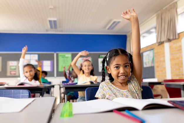 Retrato de uma menina de escola biracial feliz levantando a mão na sala de aula da escola primária