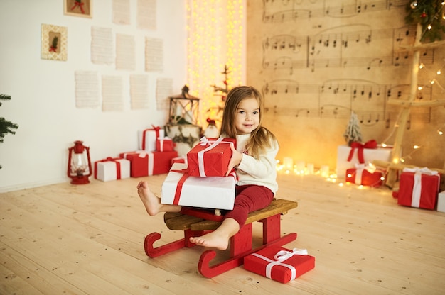 Foto retrato de uma menina em um trenó vermelho com um presente em um fundo de luzes desfocadas